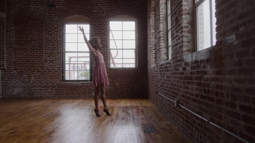 A young woman in a pink dress dances ballet in an empty room with a wooden floor and multiple sunlit windows her graceful movements highlighted by the midday light streaming in
