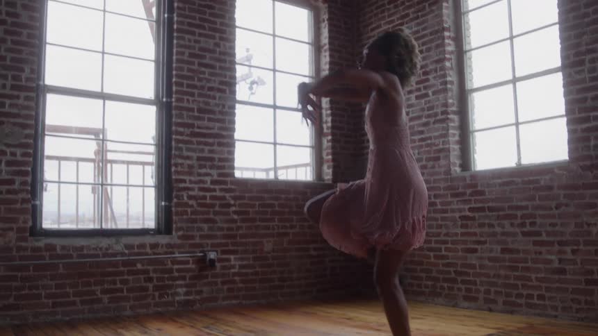 Ballerina performs grand jet with arms raised and legs extended in graceful leap before windows in industrial setting
