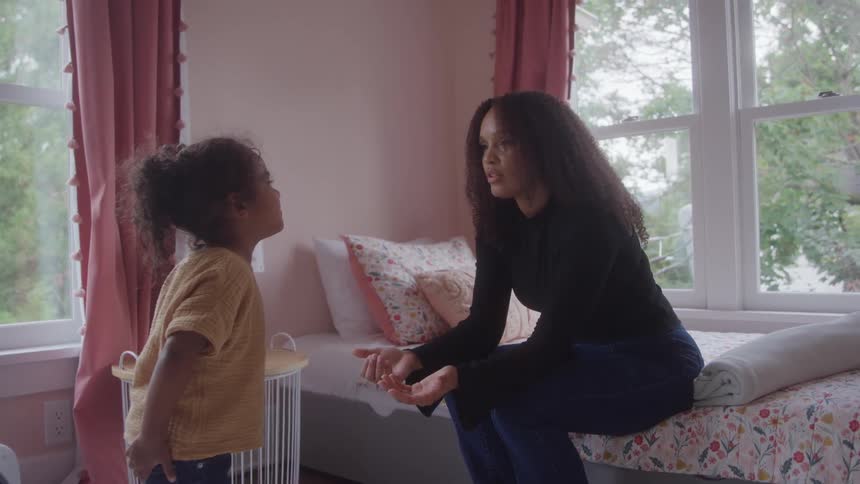 Black woman sitting on a bed trying to comfort a black female toddler who is nervously standing in front of her indoors during day time