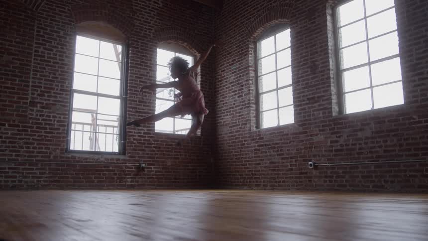A female ballet dancer performs gracefully in an empty room with a wooden floor and exposed brick walls at midday her movements illuminated by soft natural light