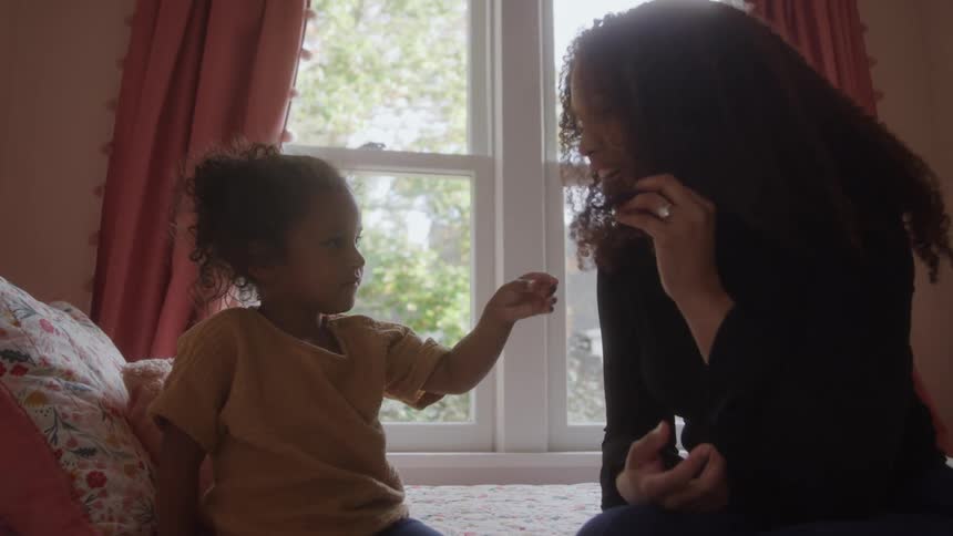 Child touches her mothers hair as they smile while sitting on the bed