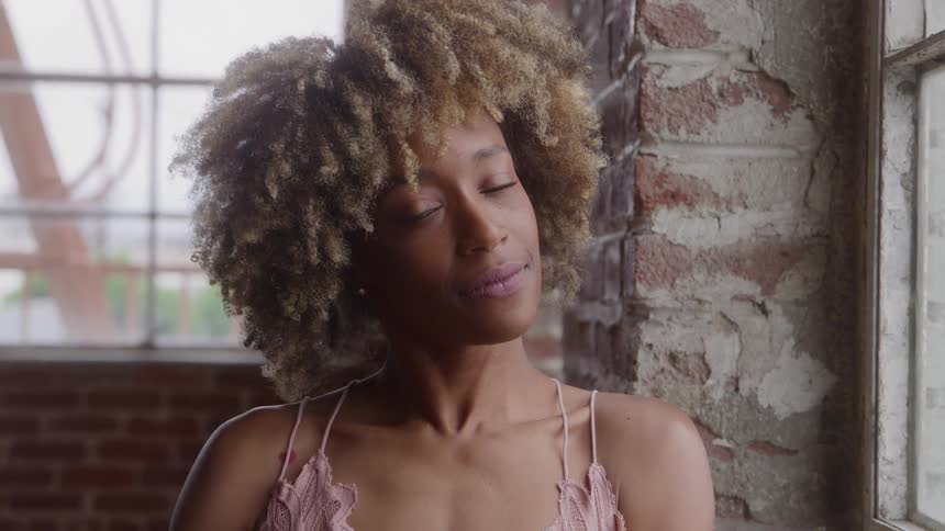 Woman with voluminous curly hair faces the camera beside a brick wall with peeling paint