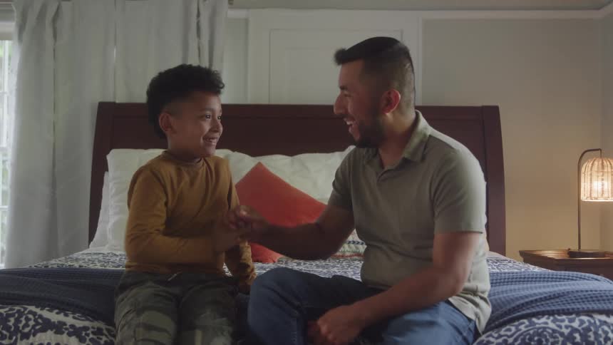 Young boy and young man sitting on a bed and high fiving while smiling indoors during day time