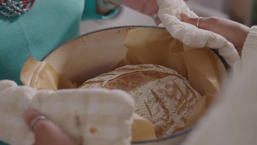 The hands of a woman hold a large gray dutch oven containing a freshly baked loaf of sourdough bread lined with brown parchment paper