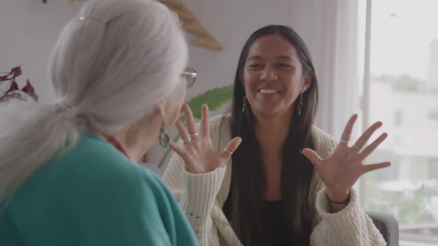 Woman in light sweater gestures smiling toward elderly whitehaired woman with glasses and turquoise top in room with plant and sheer curtain