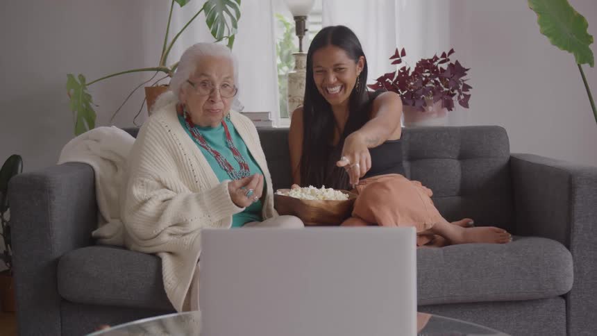 Two women laugh while watching a laptop together on the couch