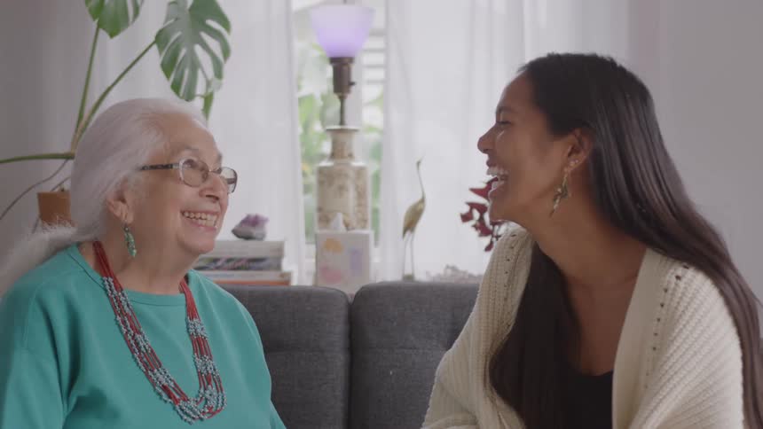 Two women sit on a gray couch talking in a welllit room with plants and white curtains in the background