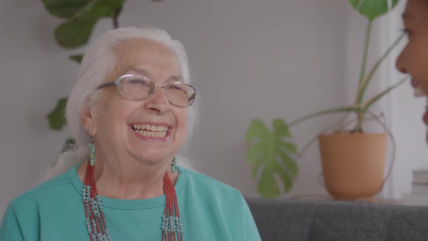 A close view of a joyful older woman with white hair and glasses wearing a teal shirt and red beaded necklaces as she smiles and laughs next to another woman in a bright living room
