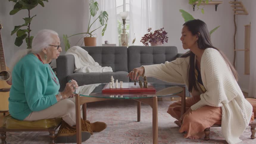 An older woman in a teal sweater and a younger woman in a cream cardigan sit across a glass table playing a game of chess in a bright plant filled living room