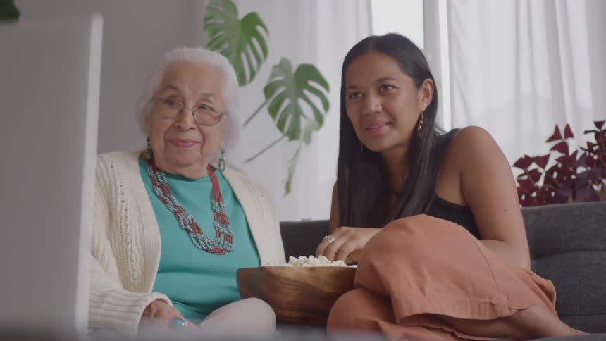 Elderly whitehaired woman with glasses and turquoise necklace sits beside woman in black top with bowl of popcorn in room with plants and sheer curtains