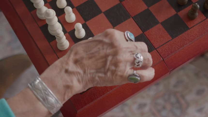 A closeup shot shows a hand wearing multiple rings and a silver cuff reaching onto a red and black chess board and moving a small white chess piece