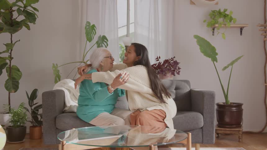 People embrace on a gray couch in a welllit room with plants and sheer curtains in the background