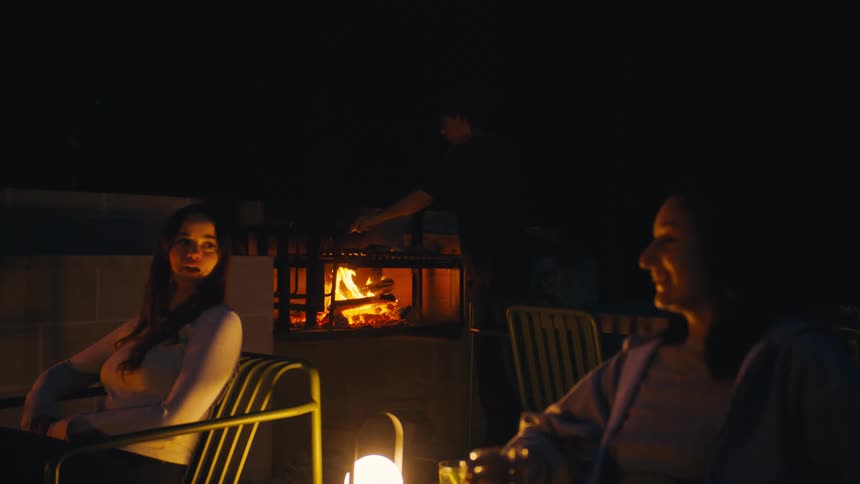 Two young women sitting down and talking while a man on the background grills food on a barbecue outdoors during night time