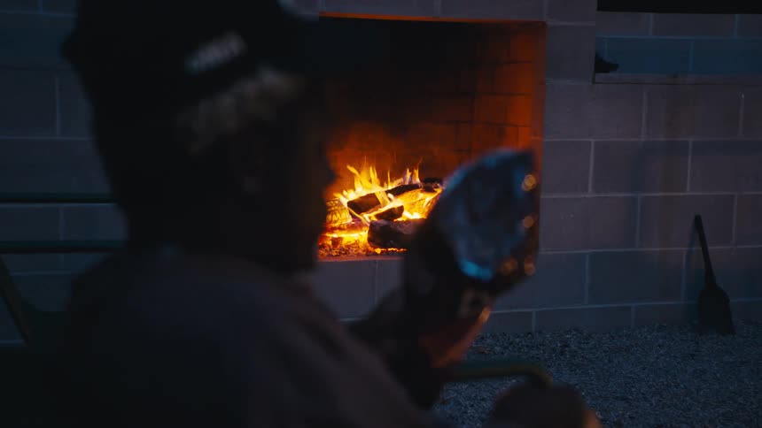 Man in dark hoodie and cap holds boot near face seated by lit fireplace in outdoor area with stone ground