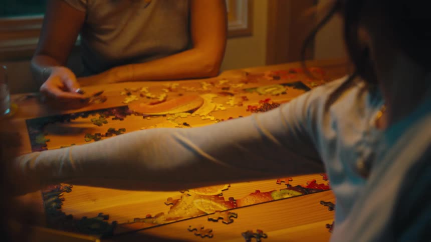 Three young women sit around the dining table at sunset playing a picture puzzle game as warm light from the dining room softly illuminates their focused faces and scattered puzzle pieces