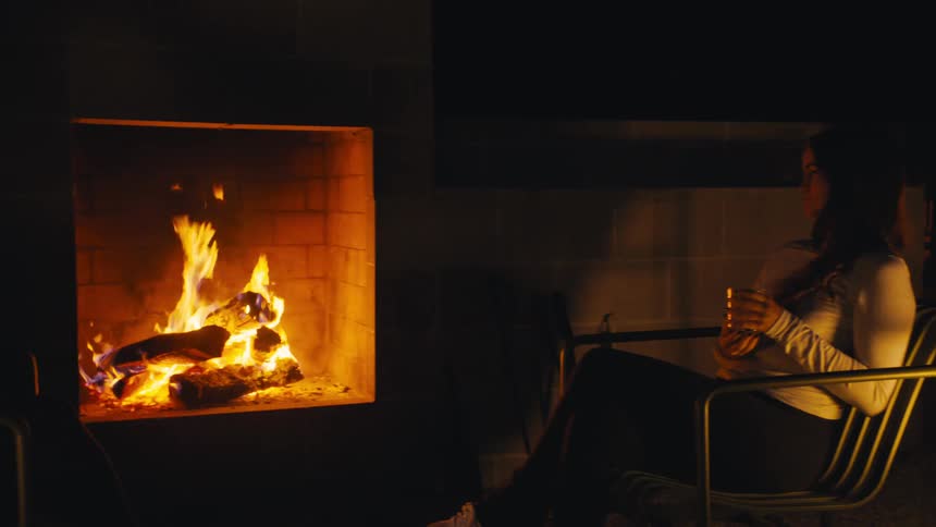 Three women sitting near a fireplace talking while a man is grilling meat on a barbecue