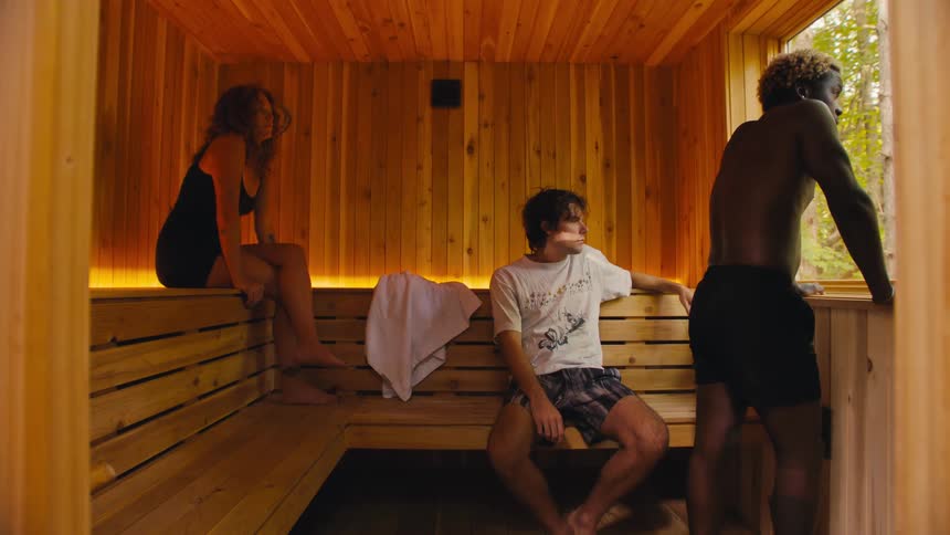 Three people a woman and two men sit on wooden benches inside a wooden room with one man standing near an open window looking outside while the other two sit and rest