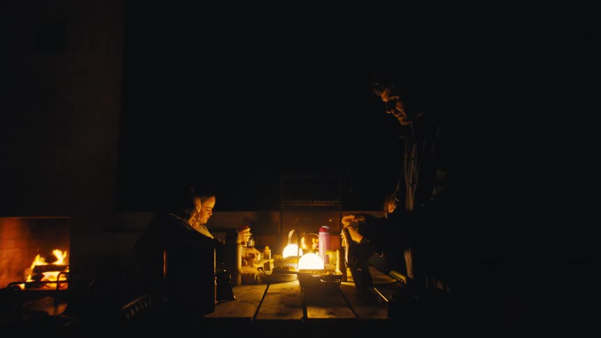 Four people talk around wooden table lit by lantern with fireplace burning in outdoor area at night