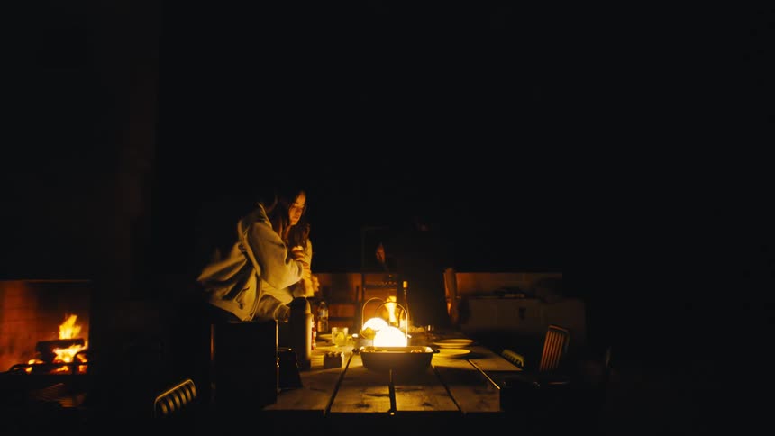 A wide shot in a dark room shows people around a wooden table with firelight from a fireplace and lanterns on the table providing the only illumination also theres one person cooking in the oven