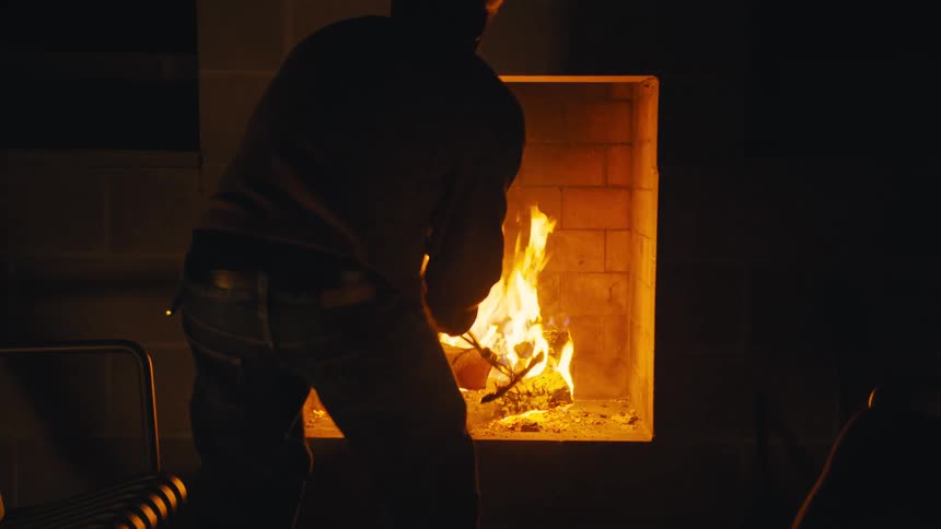 Man tends a fireplace with wood as the flames light up the dark room