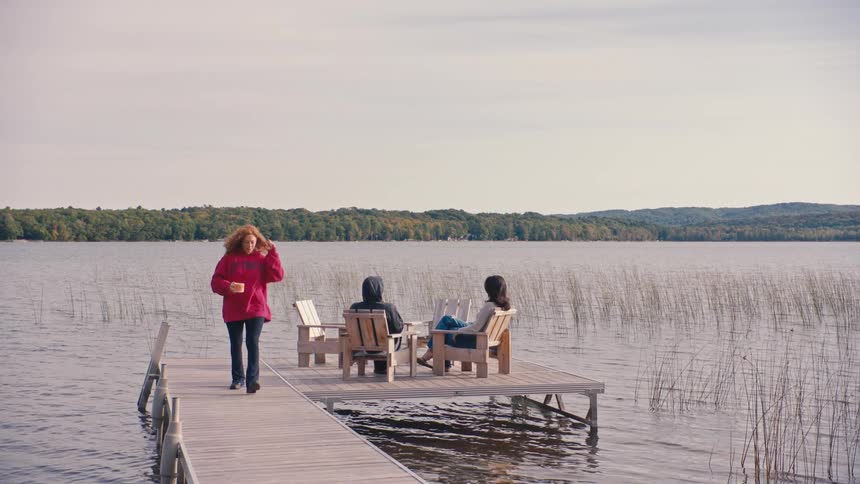 Three women sitting on chairs on a riverside deck and one of them gets up and walks away holding a cup
