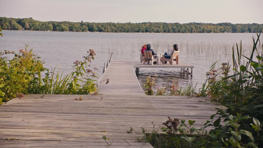 Friends sit in chairs at the end of a dock across the lake from a forest in autumn
