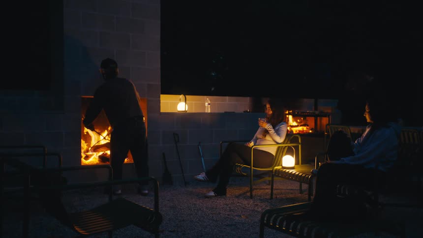 Small group of people having a barbecue outdoors with some of them tending to the fire and the food while others sit around and have drinks outdoors during night time