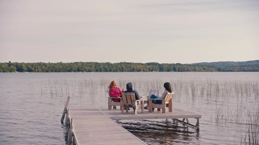 A person with long curly hair wearing a red hoodie walks down a wooden dock toward two other people sitting in chairs at the end of the dock all overlooking a wide body of water and a forested shoreline