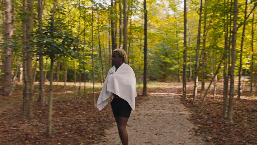 A gravel path through tall autumn trees leads into the woods as a black man walks into a brick shower building