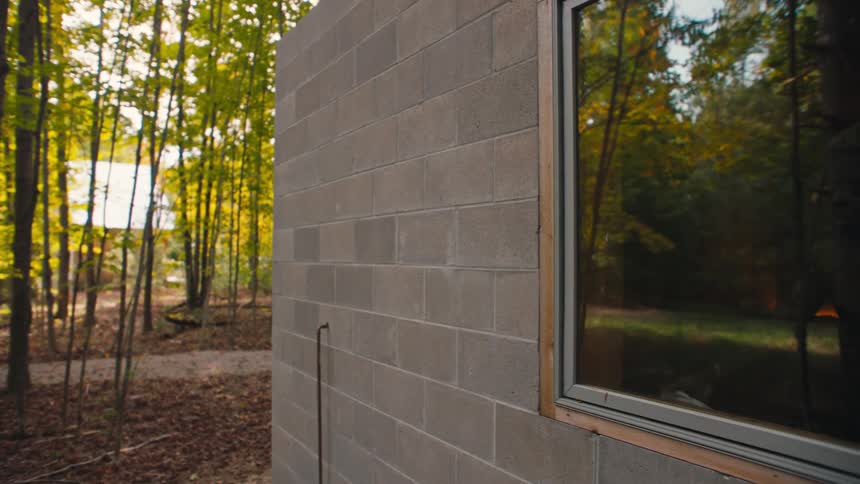 A building stands quietly in the woods surrounded by tall trees under the midday sun through one of its windows a person is visible opening the door and stepping inside their figure framed by dappled light filtering through the forest canopy