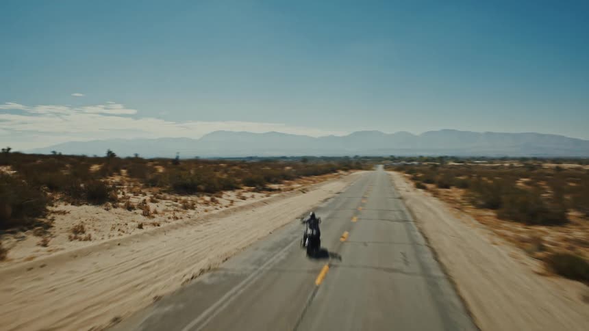 An aerial or high shot follows a motorcycle traveling on a straight paved road that extends into the distance through a desert landscape towards a distant mountain range under a clear sky