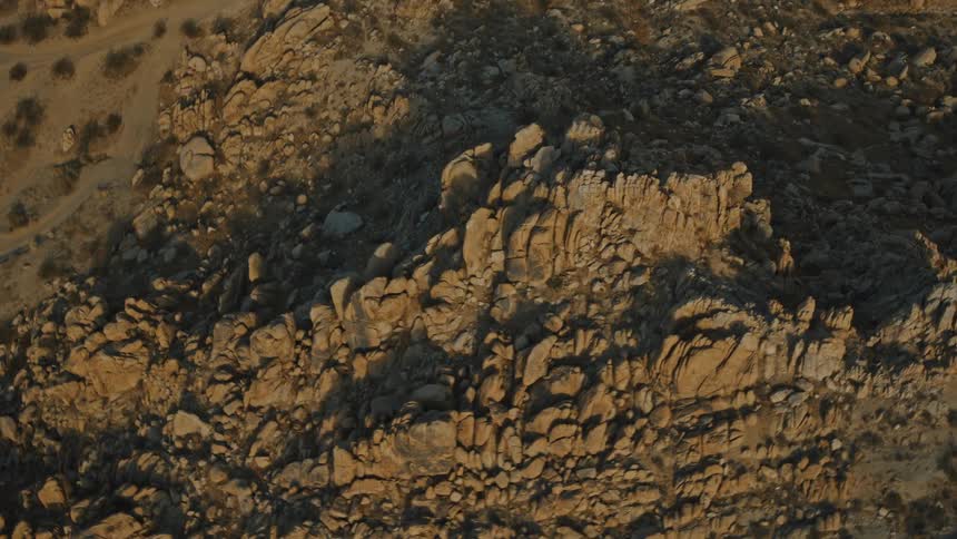 An aerial view descends slowly over a steep desert cliff composed of large sunlit brown rocks with areas of sandy ground and faint trails visible at the top