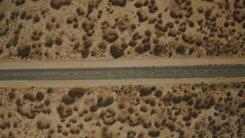 Send an elevated view looks straight down onto a long cracked asphalt road with yellow dividing lines stretching through a vast dry desert landscape with scattered brown brush