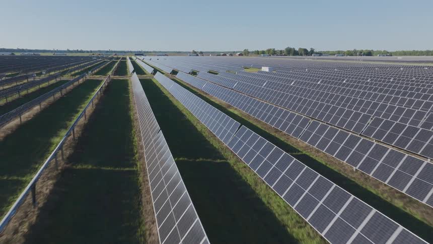A wide solar farm shows long rows of photovoltaic panels stretching across green fields under a clear sky