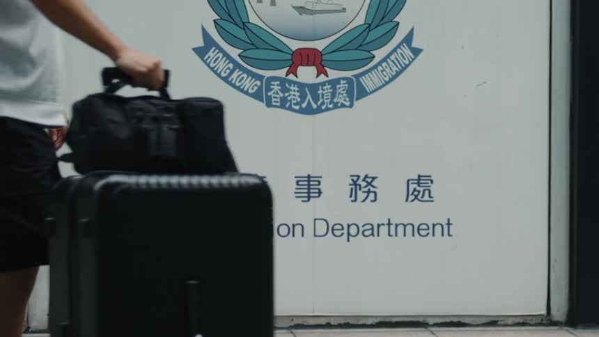 A signpost for an immigration office in hong kong written in chinese stands prominently at midday as people roll their travel briefcases past it