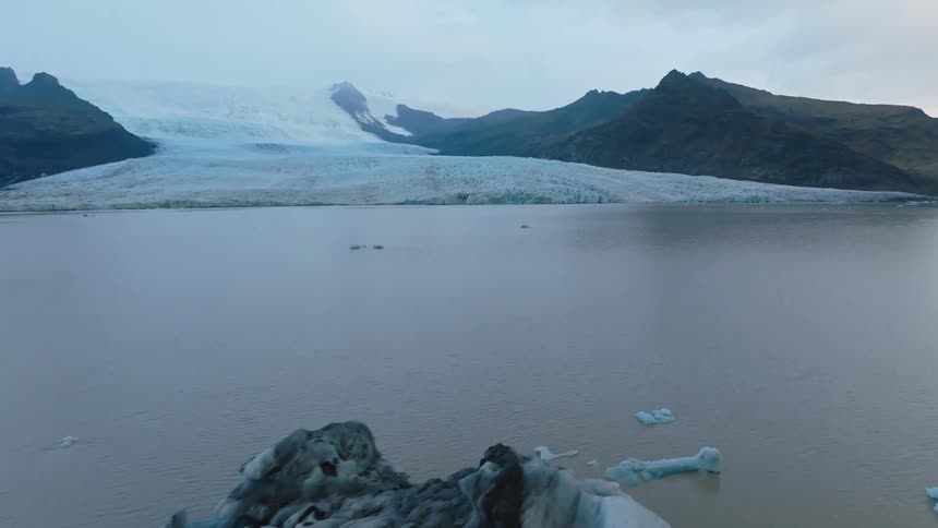 The video is a wide establishing shot of a large glacial lake with an ice sheet resting on a rocky mountain range in the background several icebergs float in the foreground including one larger piece with a distinct blue tint