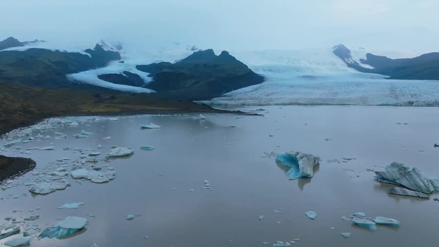 An aerial view shows large bluetinged icebergs floating in a grey glacial lagoon with a vast snowcovered glacier and dark mountains forming the background under an overcast sky