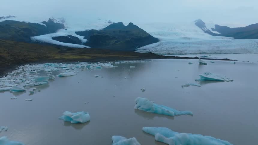 An icy lake lies still beneath a cloudy sky surrounded by vast towering mountains the muted light casts a soft silvery tone across the frozen surface while the rugged peaks rise solemnly in the distance