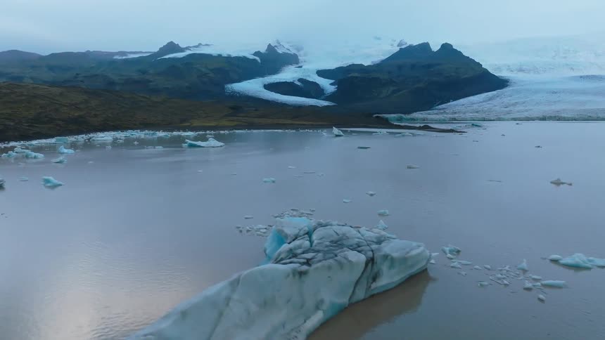 Glacier icebergs with bright blue streaks float in the gray glacial lake with dark rocky snow covered mountains and a curving white glacier filling the background under a dim sky