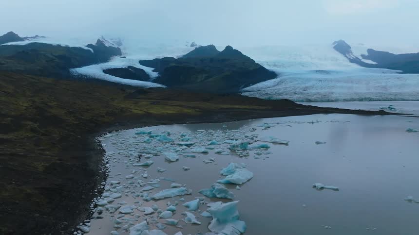 An aerial wide shot shows a large glacier and snowcovered mountains in the background and a glacier lake filled with floating icebergs