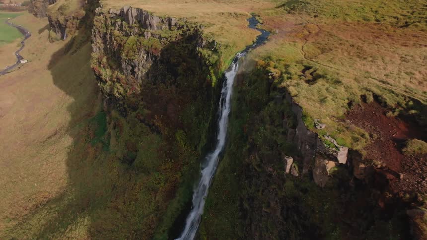 An aerial shot shows a stream of water falling down a steep cliff face creating a waterfall that lands next to a winding river in a large grasscovered landscape