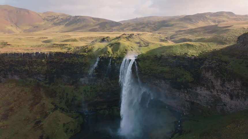 A wide sweeping view shows a large waterfall plunging down a steep mosscovered cliff that is part of a long mountain range under a cloudy sky