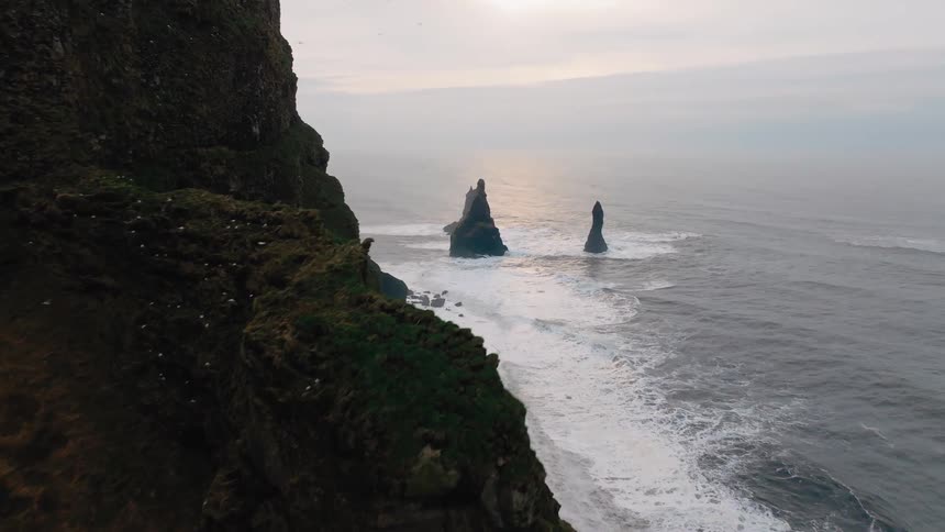Rock formations rise from the sea beside vegetationcovered cliffs under a cloudy sky