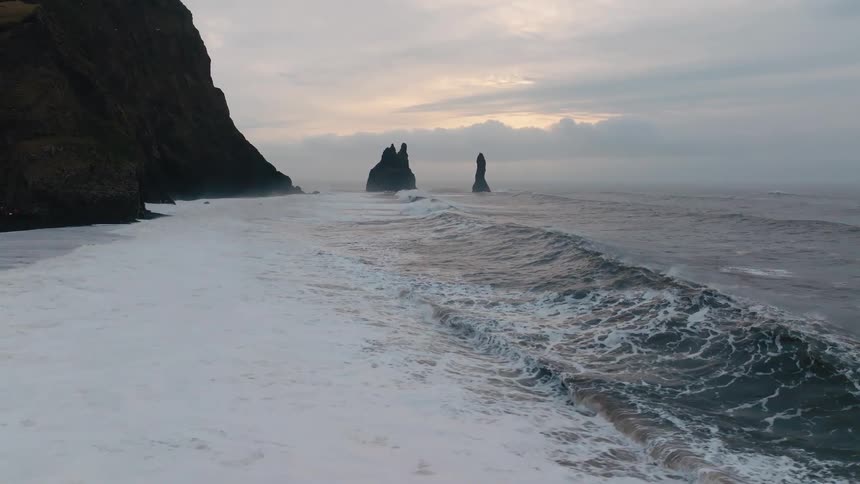 Send high ocean waves with thick white foam surge toward a black sand beach and a dark cliff face with tall basalt sea stacks visible offshore under a cloudy pale sky