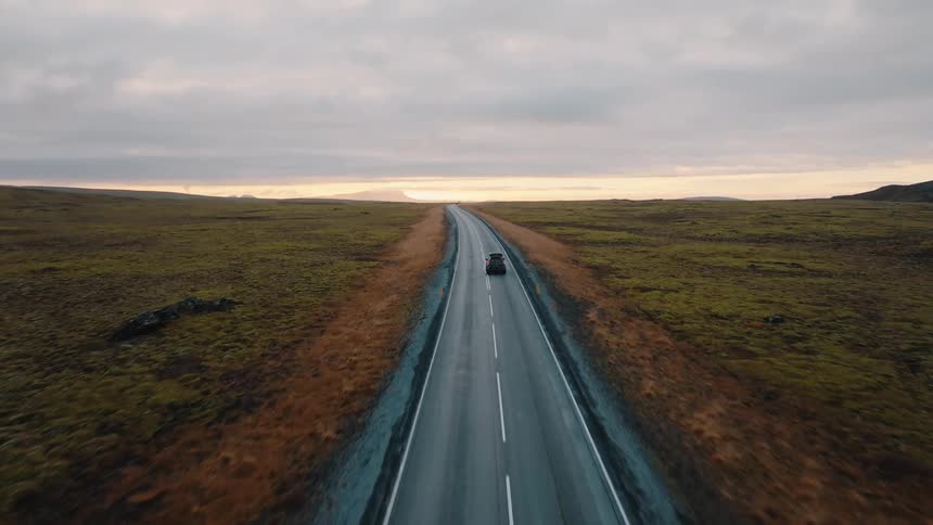 An elevated perspective follows a black car with a rooftop tent driving away on a long straight asphalt road surrounded by rolling brown and green mossy terrain under a pale sunset sky