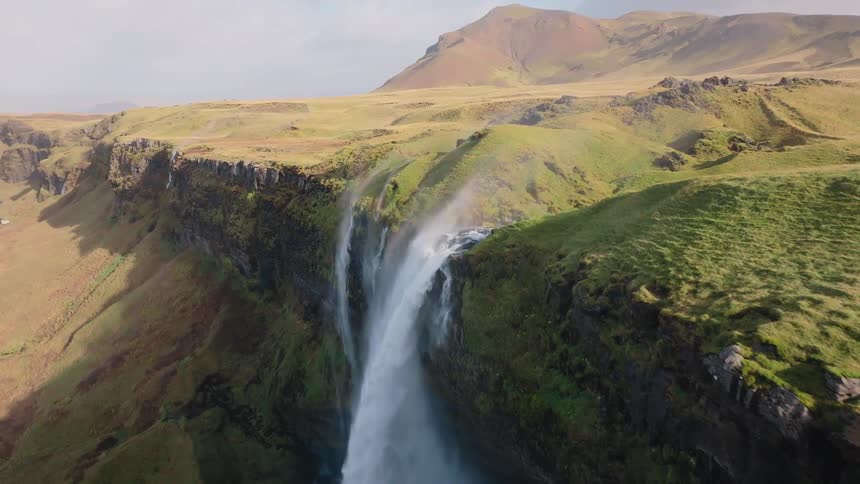 A waterfall cascades down a mountain blanketed in short grasses glistening under the bright sun on a clear day