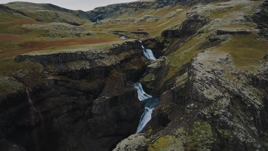The video is an aerial view of a canyon landscape dominated by shades of green and brown with rugged mossy cliffs a waterfall cascades down the dark rock face into the gorge below with the river visible flowing in the distance