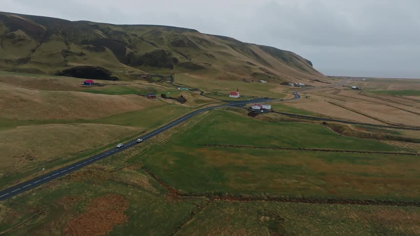 Send an elevated view shows a black asphalt road winding through rolling green and brown hills with scattered white houses and a small church near the ocean under an overcast sky
