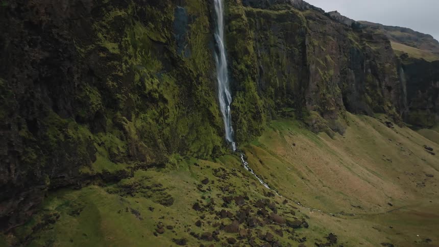 An aerial dolly in view captures a tall narrow waterfall plunging down the side of a massive dark cliff face covered in bright green moss and scrub leading to a rocky expansive valley floor under an overcast sky