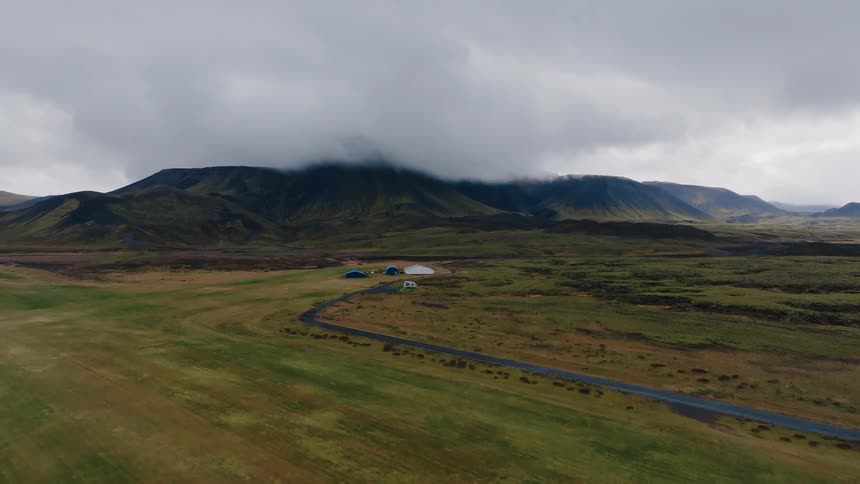 Isolated houses appear at the end of a winding road in open terrain with cloudcovered mountains in the background